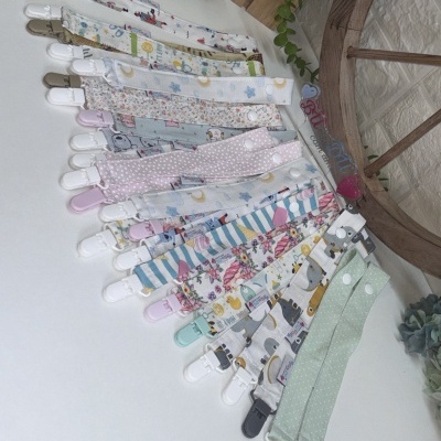 Presilhas para chupetas em várias estampas e cores sobre mesa branca com plantas e decoração ao fundo.