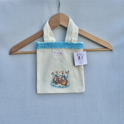 Saco de pano creme com alças largas e estampado de barco com animais, pendurado num cabide de madeira.