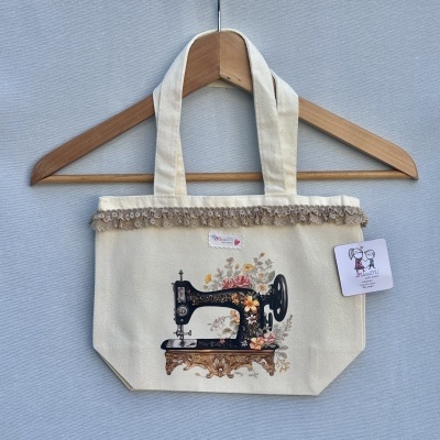 Mala de tecido crua com imagem de máquina de costura e flores coloridas, com renda bege e etiqueta pendurada