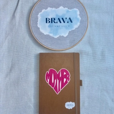 Caderno castanho com autocolante MOTHER e bastidor de madeira com tecido cinza e logotipo BRAVA ESTAMPAGEM