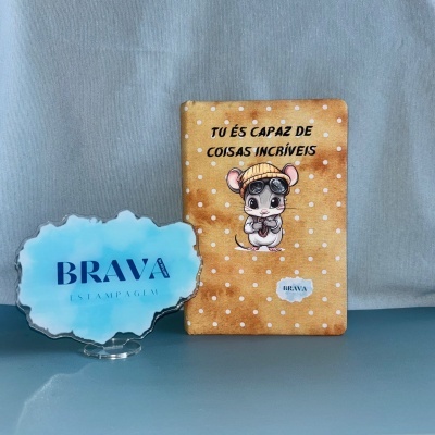 Caderno com capa castanha e ilustração de rato com gorro e óculos, placa azul translúcida com texto BRAVA ESTAMPAGEM.