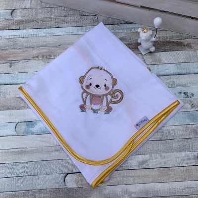 Manta branca com bordas amarelas e bordado de macaco, sobre madeira, com estatueta ao lado