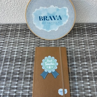 Caderno castanho com selo azul Best Teacher em superfície de vime e bastidor de tecido azul claro