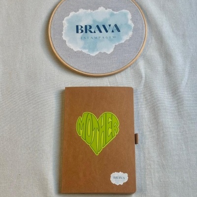 Caderno castanho com coração verde e bastidor de bordado com logótipo BRAVA ESTAMPAGEM sobre tecido claro