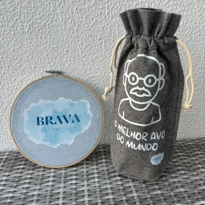 Saco cinzento de tecido com desenho de rosto e texto O MELHOR AVÔ DO MUNDO, e bastidor de madeira com tecido azul claro com texto BRAVA ESTAMPAGEM