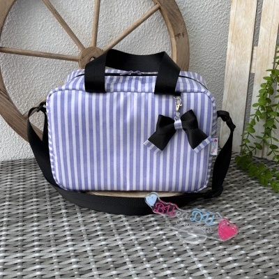 Mala riscada branca e lilás com alças pretas e laço, pendente colorido Bia Day, sobre mesa cinzenta com roda de madeira ao fundo