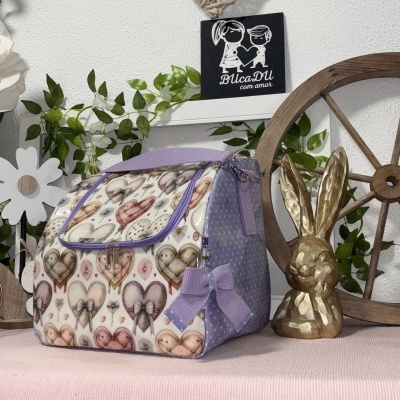 Bolsa quadrada lilás com padrão de corações em tecido, decorada sobre mesa rosa com elementos decorativos