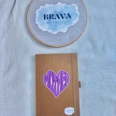 Caderno bege com autocolante MOTHER sobre tecido azul e bastidor com texto BRAVA ESTAMPAGEM