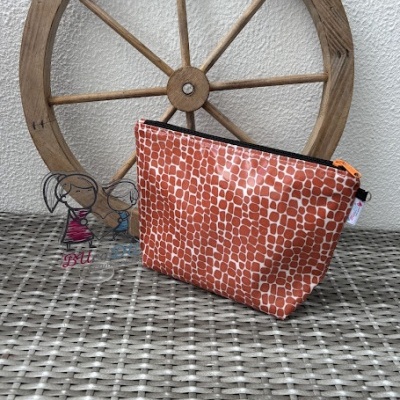 Necessaire triangular vermelha com padrão de escamas e fecho laranja, sobre mesa de rattan, com roda de madeira ao fundo e figuras acrílicas de crianças.