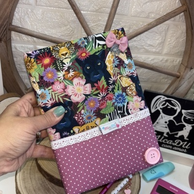 Capa de tecido para livro com padrão de animais e flores coloridas e parte inferior rosa com bolinhas brancas e botão.