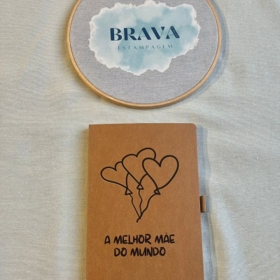 Caderno castanho com balões em forma de coração e texto A MELHOR MÃE DO MUNDO, e bastidor de madeira com pano cinzento e texto BRAVA ESTAMPAGEM