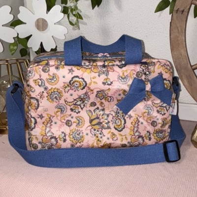 Mala retangular floral rosa com alças azuis sobre tecido rosa claro.