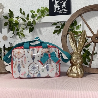Bolsa branca decorada com filtros dos sonhos coloridos, alça azul, laço e detalhes vermelhos, sobre tecido rosa com decoração ao fundo.
