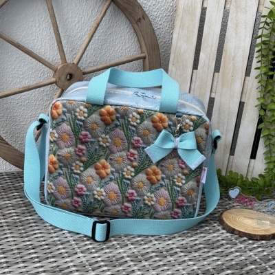 Mala azul claro bordada com flores coloridas e laço, colocada sobre superfície cinza entre roda e palete de madeira