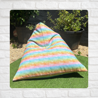 Puff triangular colorido com riscas pastel em relva artificial e vasos de plantas ao fundo