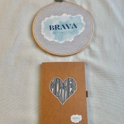 Bordado redondo e caderno castanho com texto MOTHER e BRAVA