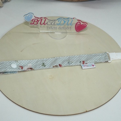 Fita com padrão de bolinhas e flores e clip branco sobre superfície redonda de madeira com suporte acrílico escrito BUcaDU com amor