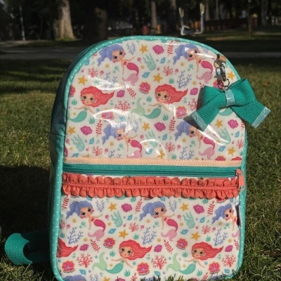 Mochila infantil branca com padrão de sereias e laço verde à frente, num parque.