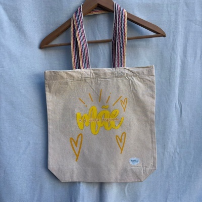 Saco de tecido bege com texto amarelo e alças listradas coloridas pendurado num cabide