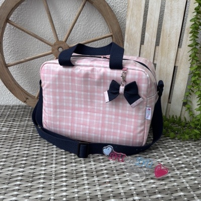 Mala rosa com padrão xadrez e alças negras sobre superfície trançada com roda de madeira ao fundo.