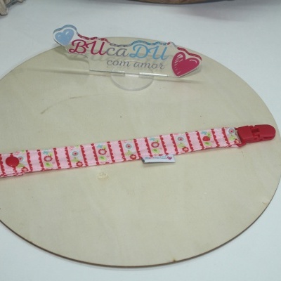 Fita para chupeta rosa com padrão colorido e fecho vermelho sobre suporte de madeira