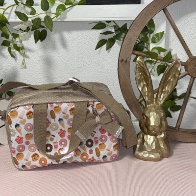 Mala pequena com padrão de doces, alças bege, figura de coelho dourado, roda de madeira e placa preta com texto.