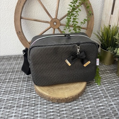 Bolsa quadrada preta com padrão quadriculado branco e laço preto pendurado no zíper, sobre base de madeira com decoração de roda de madeira e plantas.