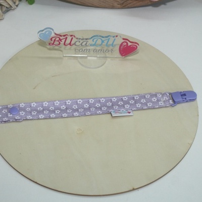 Pulseira lilás com padrão de patas brancas e fecho plástico sobre base de madeira clara com suporte acrílico com texto BUcaDU