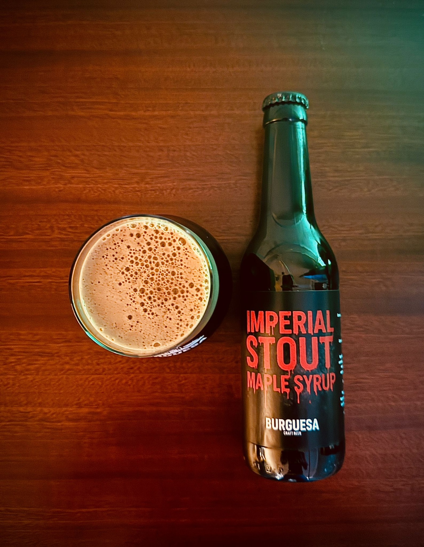 Garrafa de cerveja artesanal escura com rótulo Imperial Stout Maple Syrup Burguesa e copo com cerveja foam em mesa de madeira.
