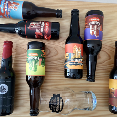 Garrafas de cerveja BURGUESA coloridas e um copo transparente sobre uma mesa de madeira