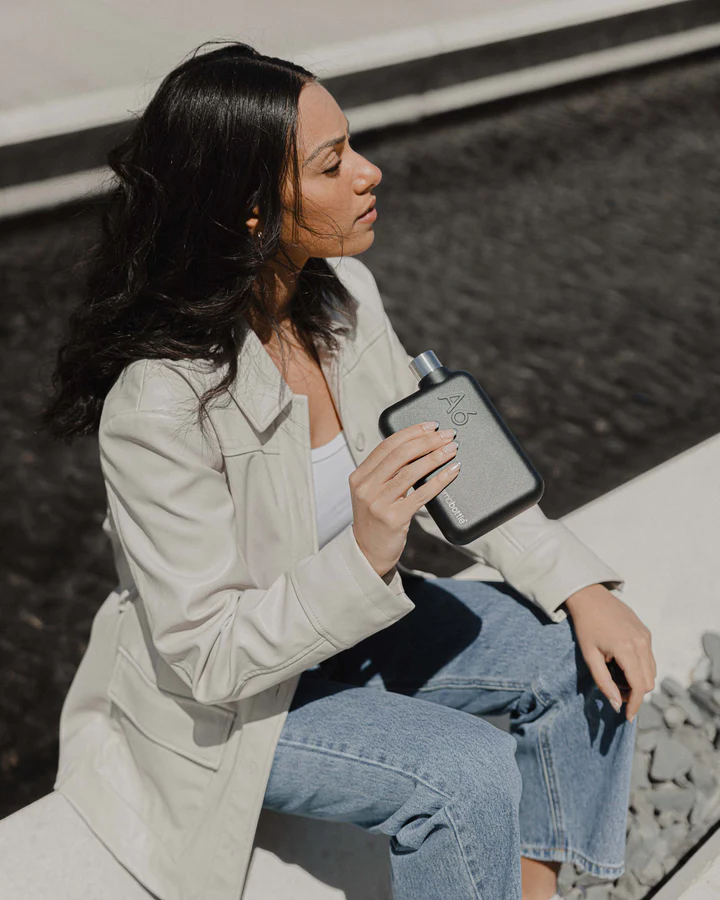 A6 memobottle Stainless Steel - BLACK Mulher segurando frasco preto com símbolo de porcentagem sentada junto a água e pavimento branco