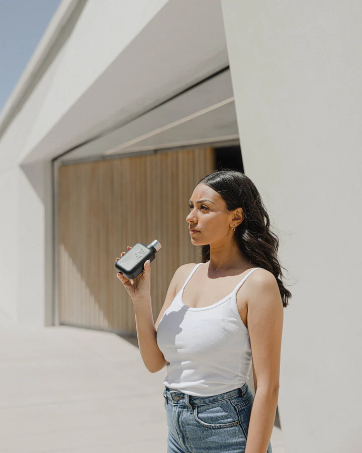 A7 memobottle Stainless Steel - BLACK Mulher segurando dispositivo eletrônico ao ar livre com parede branca e porta de madeira ao fundo.