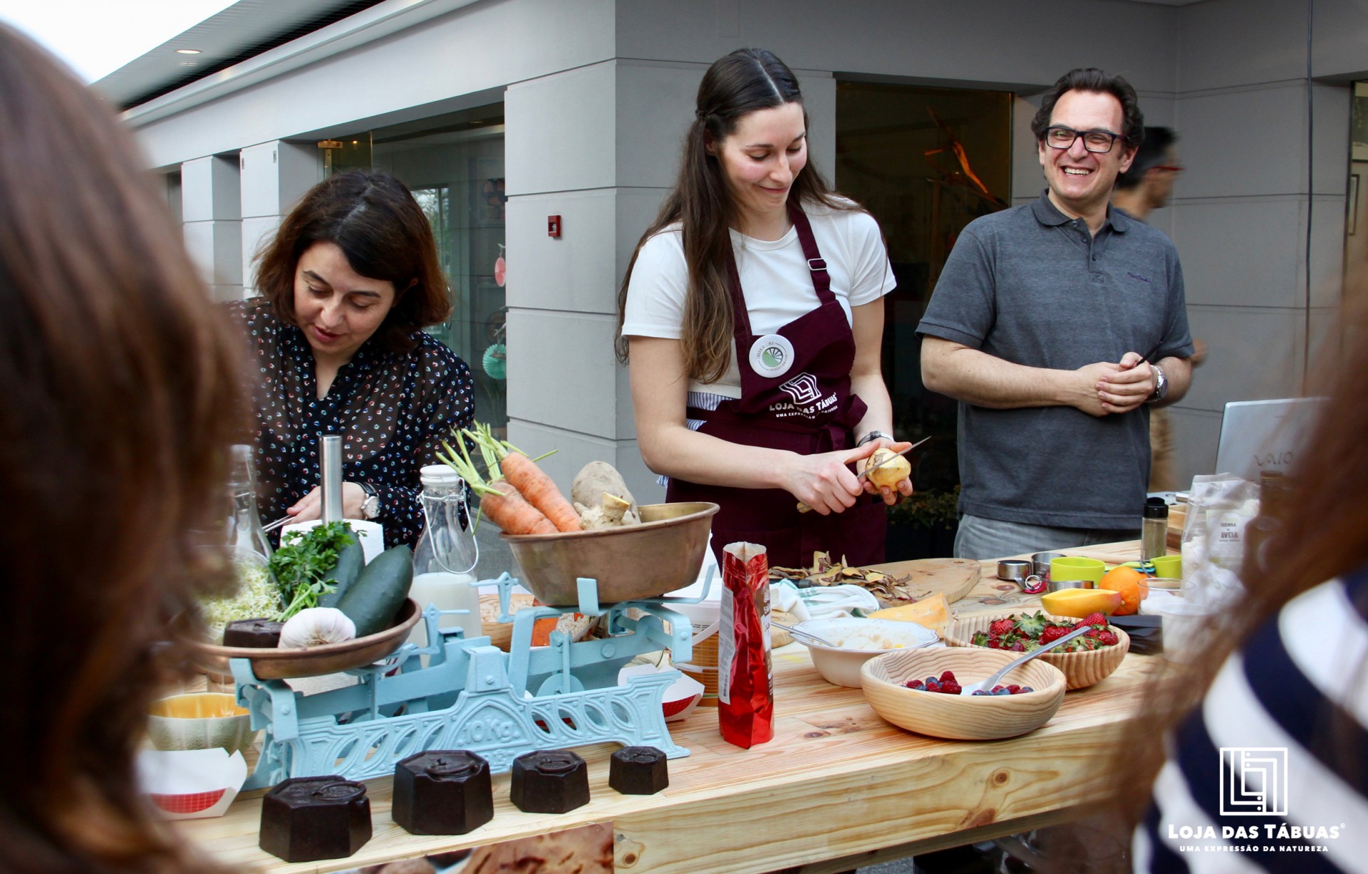 Mesa com frutas, legumes e utensílios de cozinha com pessoas ao redor
