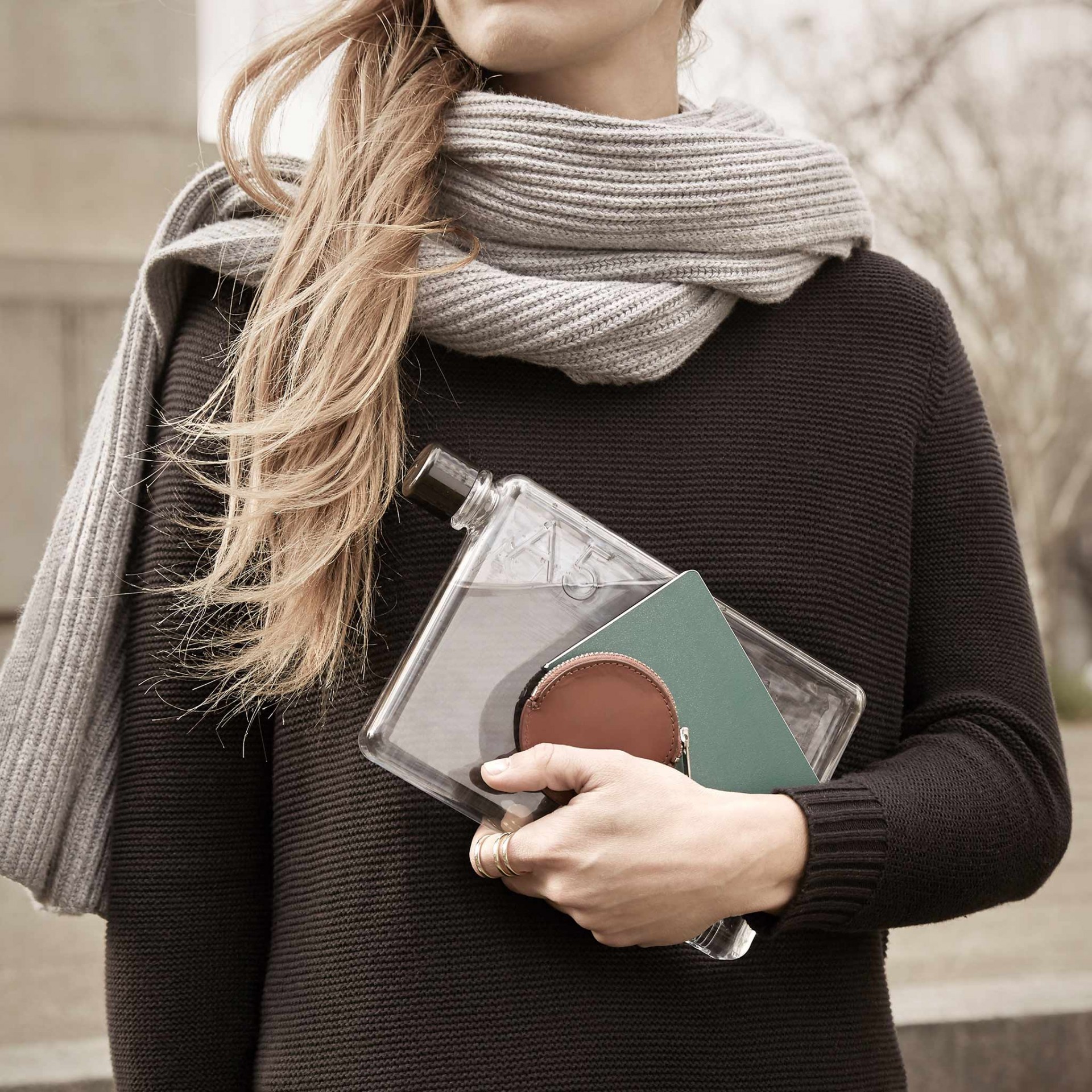 A5 memobottle Mulher segurando garrafa transparente, carteira castanha e objeto verde, com vestido preto e cachecol cinzento