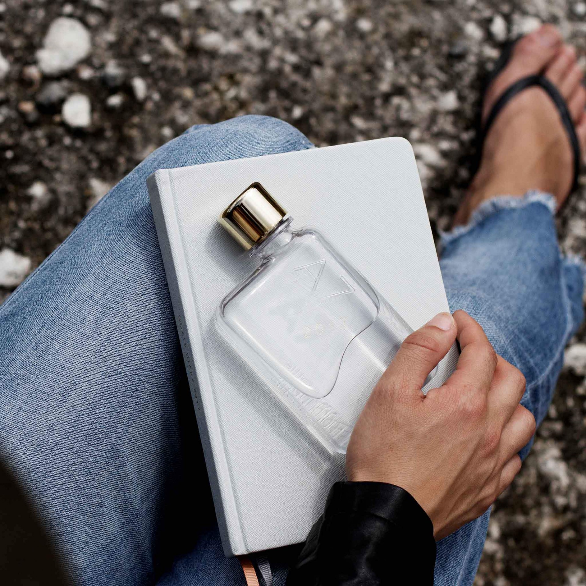 A7 memobottle Frasco de perfume transparente com tampa dourada sobre caderno branco na mão, fundo de pedras e perna com jeans e chinelo.