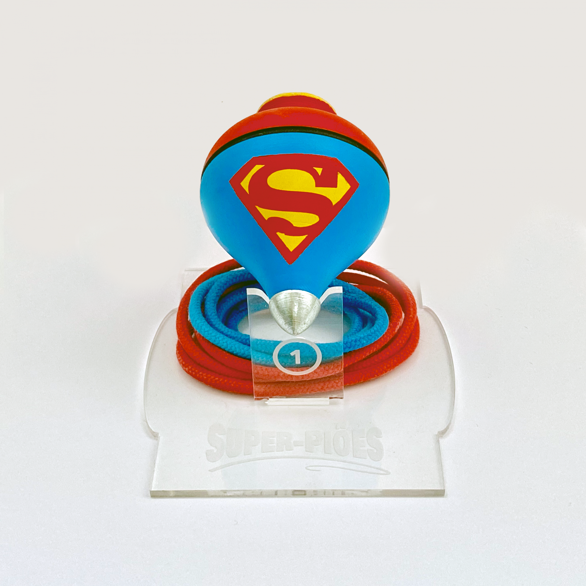 Super-Piões de Madeira Pintados à Mão SUPERMAN – Clássicos da Infância Reinterpretados por Damião Vieira Pião de madeira azul com símbolo do Superman e corda azul e vermelha numa base acrílica