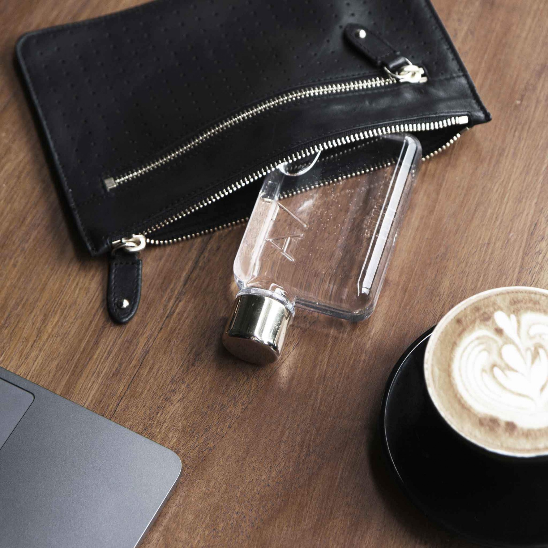 A7 memobottle Saco de mão preto em pele com fecho metálico aberto, garrafa plástica transparente, chávena preta com café e espuma, canto de portátil cinzento sobre mesa de madeira