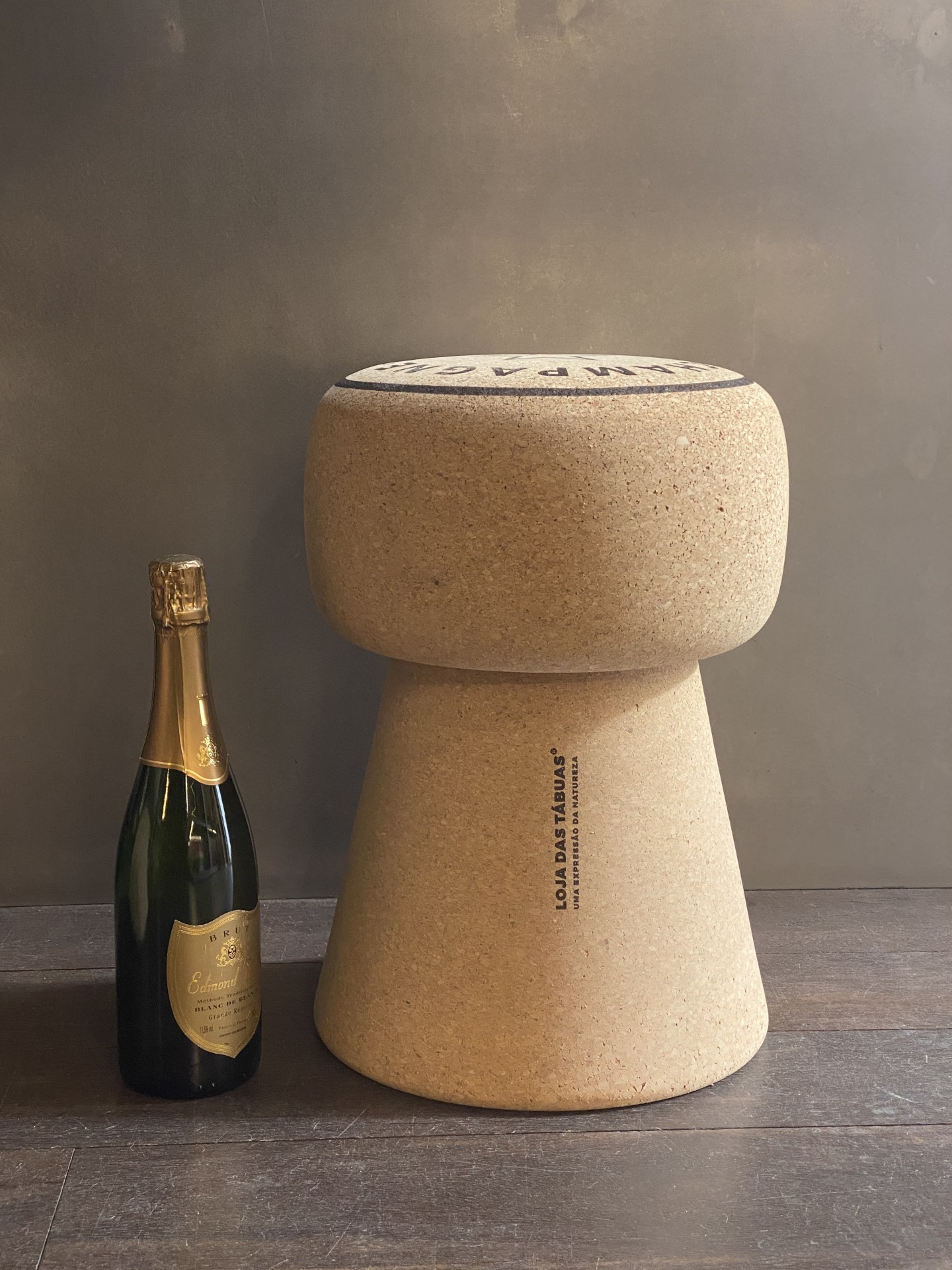 Banco em Cortiça Maciça em Formato de Rolha de Champanhe - Inscrição "Champagne" no topo Banqueta de cortiça ao lado de garrafa de champanhe sobre mesa de madeira