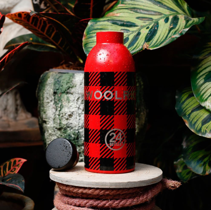 Clima Bottle - Woolrich 500ml Garrafa térmica vermelha e preta xadrez com texto WOOLRICH e logo 24 BOTTLES, sobre suporte de madeira e corda com plantas ao fundo
