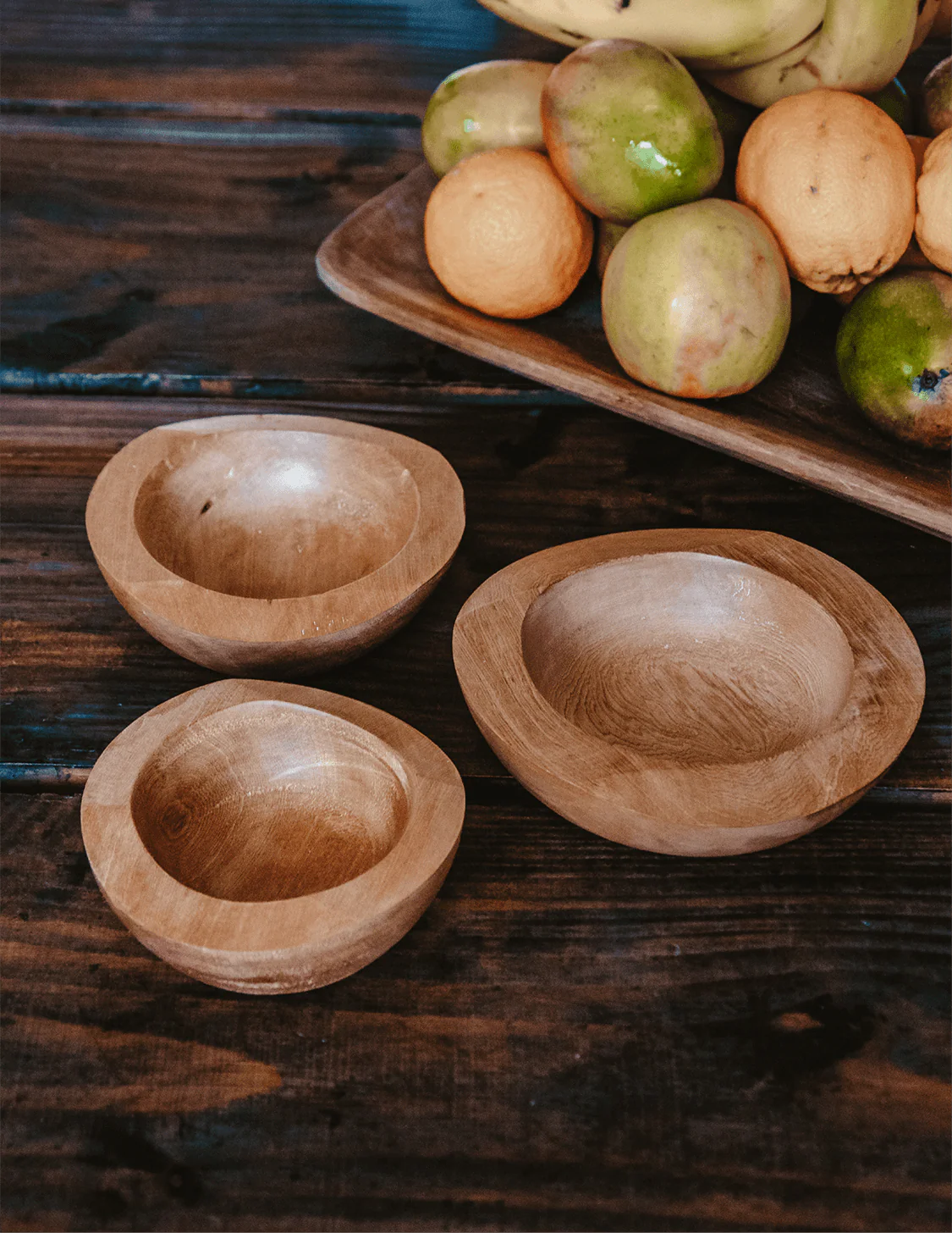 Pack IWE – Conjunto de 3 Taças em Madeira de Mañío Três tigelas de madeira natural sobre mesa escura com frutas em tabuleiro de madeira ao fundo.