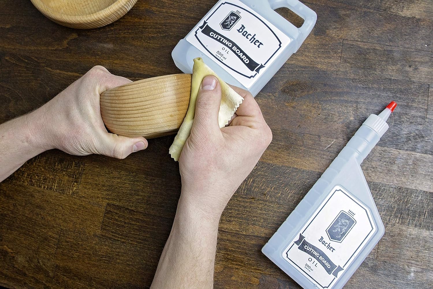 Óleo para tábuas de corte e bancadas de cozinha 200 ml Mãos limpando recipiente de madeira clara com pano, ao lado de frascos Bocher de óleo e limpador em mesa de madeira escura