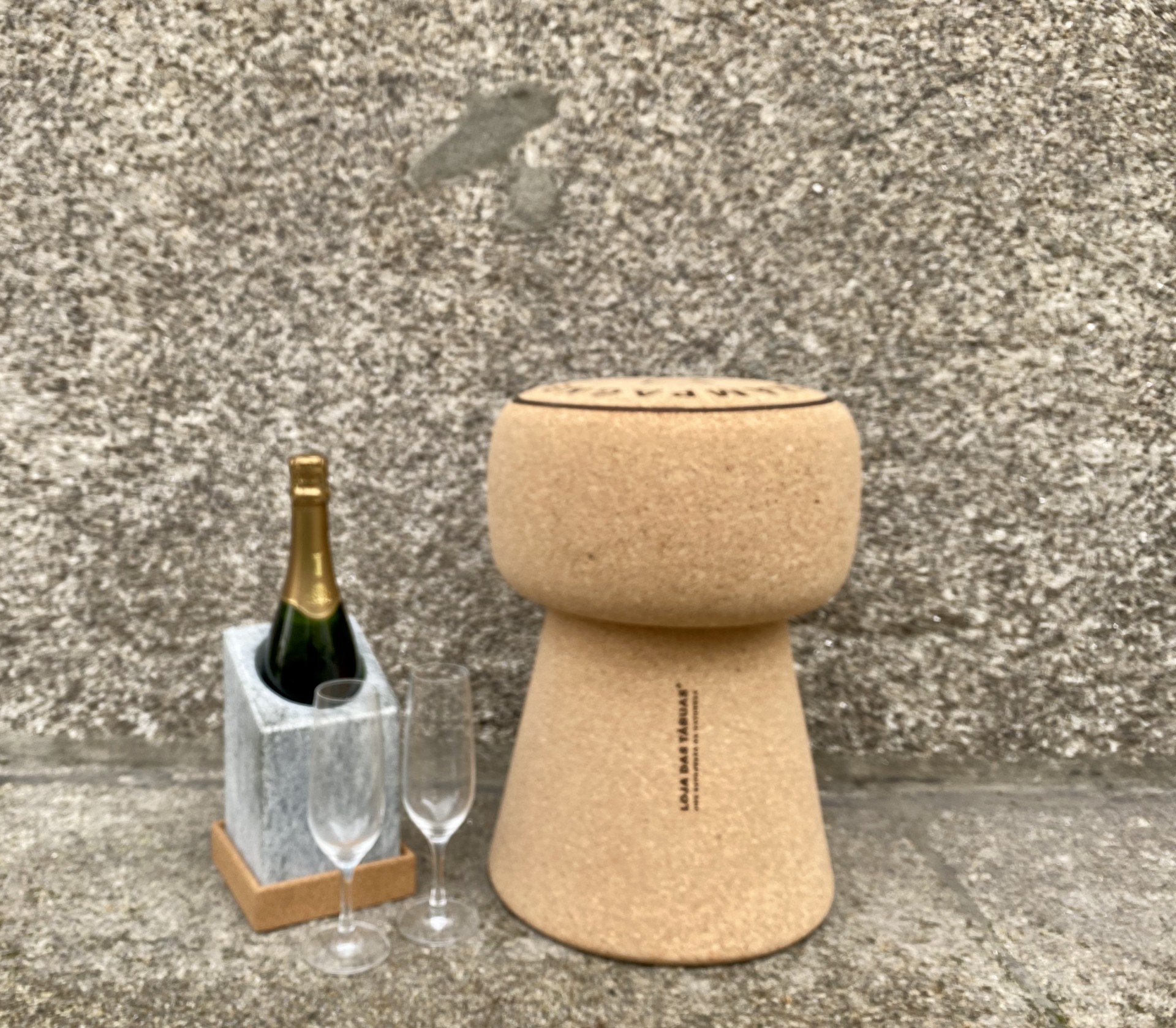 Banco em Cortiça Maciça em Formato de Rolha de Champanhe - Inscrição "Champagne" no topo Rolha de cortiça para champanhe ao lado de garrafa e copos sobre superfície de pedra