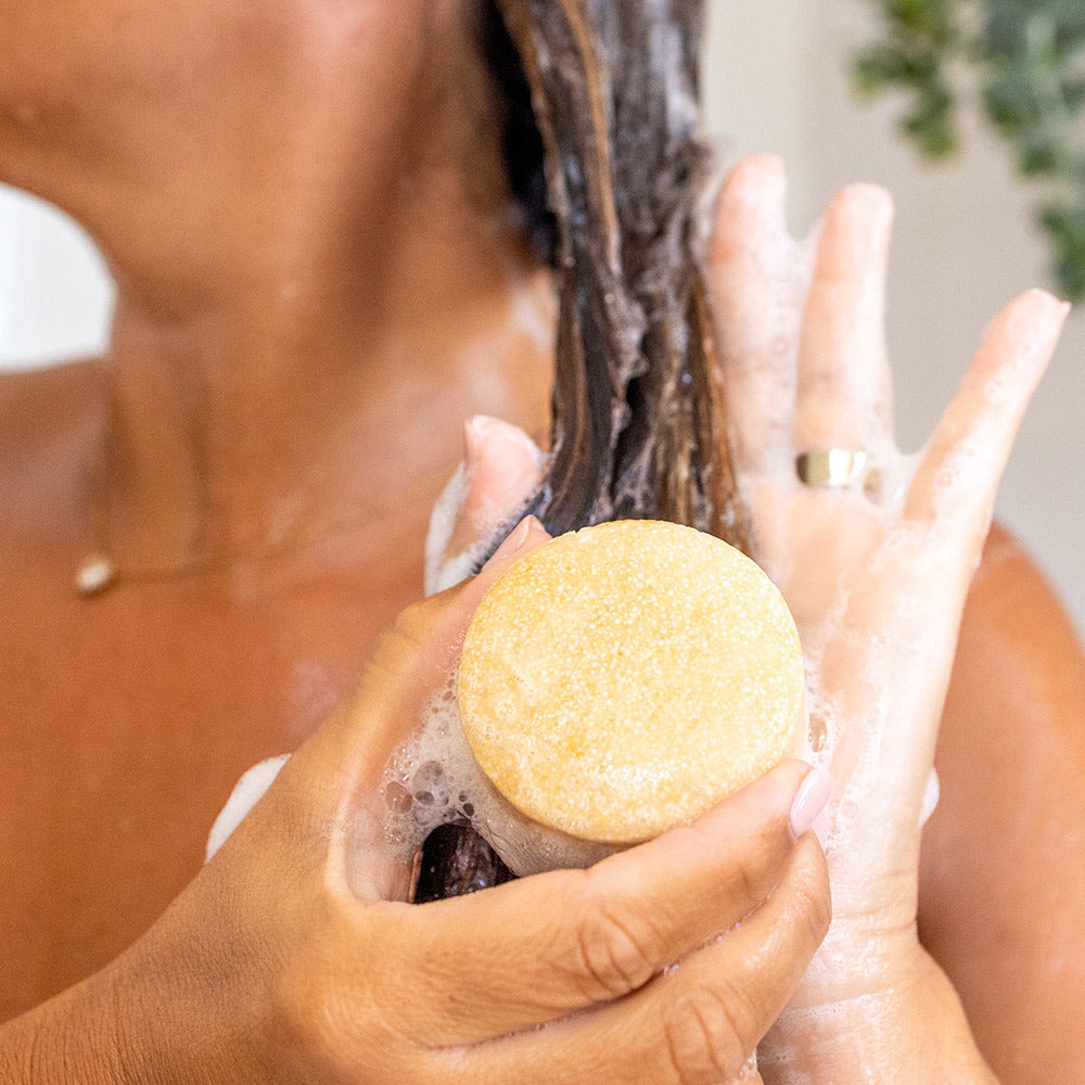 Champô Sólido NAUA Couro Cabeludo Sensível Mãos femininas aplicando shampoo sólido amarelo em cabelo molhado com espuma