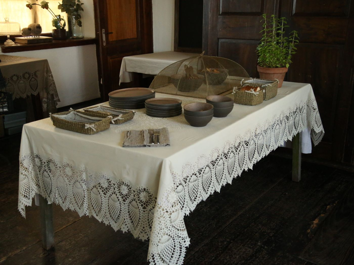 Cobertura alimentar em rede abaca 48x66x25cm - verde linho Mesa com toalha branca de renda, pratos e tigelas castanhas, cestos de vime, rede metálica para alimentos e vaso de planta