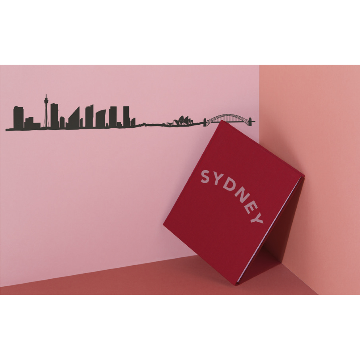 theLine® Small 50 cm - Black _ Sydney Caixa vermelha com a palavra SYDNEY apoiada num canto com parede rosa e chão vermelho, silhueta preta de Sydney na parede