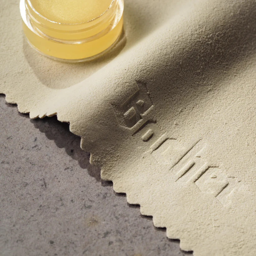 Faca Santoku Bacher em Aço Damasco com Pano de Limpeza e Cera Protetora Pano de microfibra bege com texto em relevo e recipiente de vidro com líquido amarelo