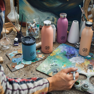 Garrafas térmicas coloridas 24Bottles em mesa de trabalho de artista com paletas e pincéis