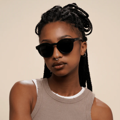 Mulher com óculos de sol de armação preta e lentes escuras, com cabelo trançado e camiseta cinzenta