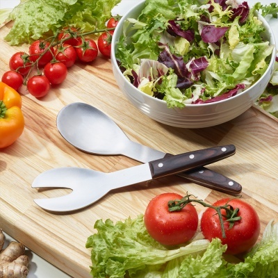 Talheres para salada com cabos de madeira numa tábua de cortar rodeada por vegetais frescos.