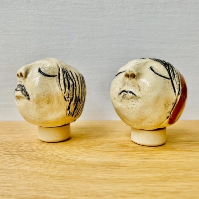 Duas esculturas cerâmicas de cabeças humanas com olhos fechados, em tons bege, sobre madeira clara